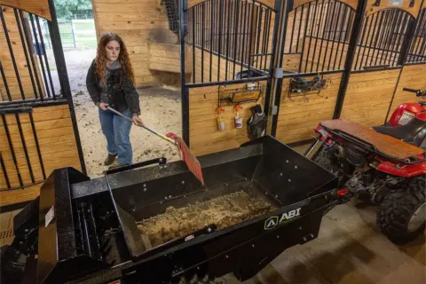 Woman mucking a stall in a barn to fill an ABI Attachments 25 Cubic Foot Ground Drive Manure Spreader